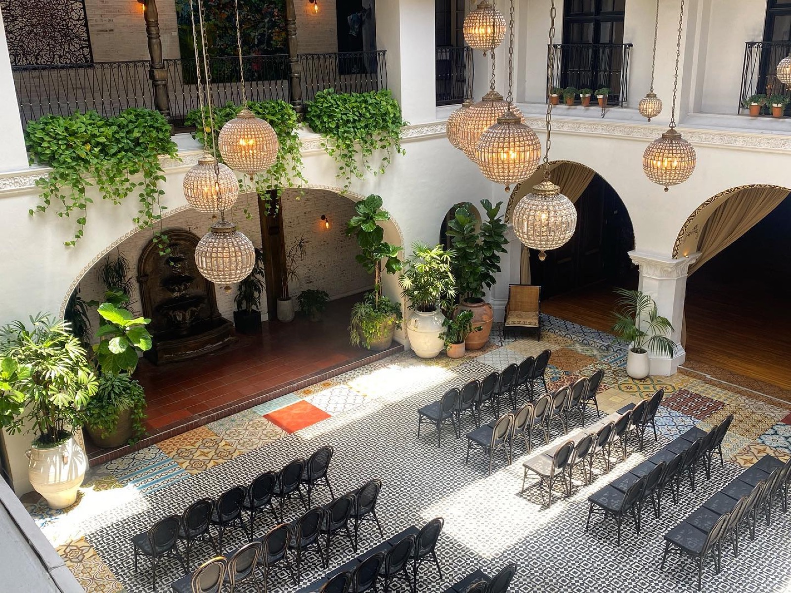 Sunlit Spanish-style courtyard with trailing pothos on the balconies, crystal globe chandeliers, potted palms and fiddle-leaf figs above a patterned tile floor